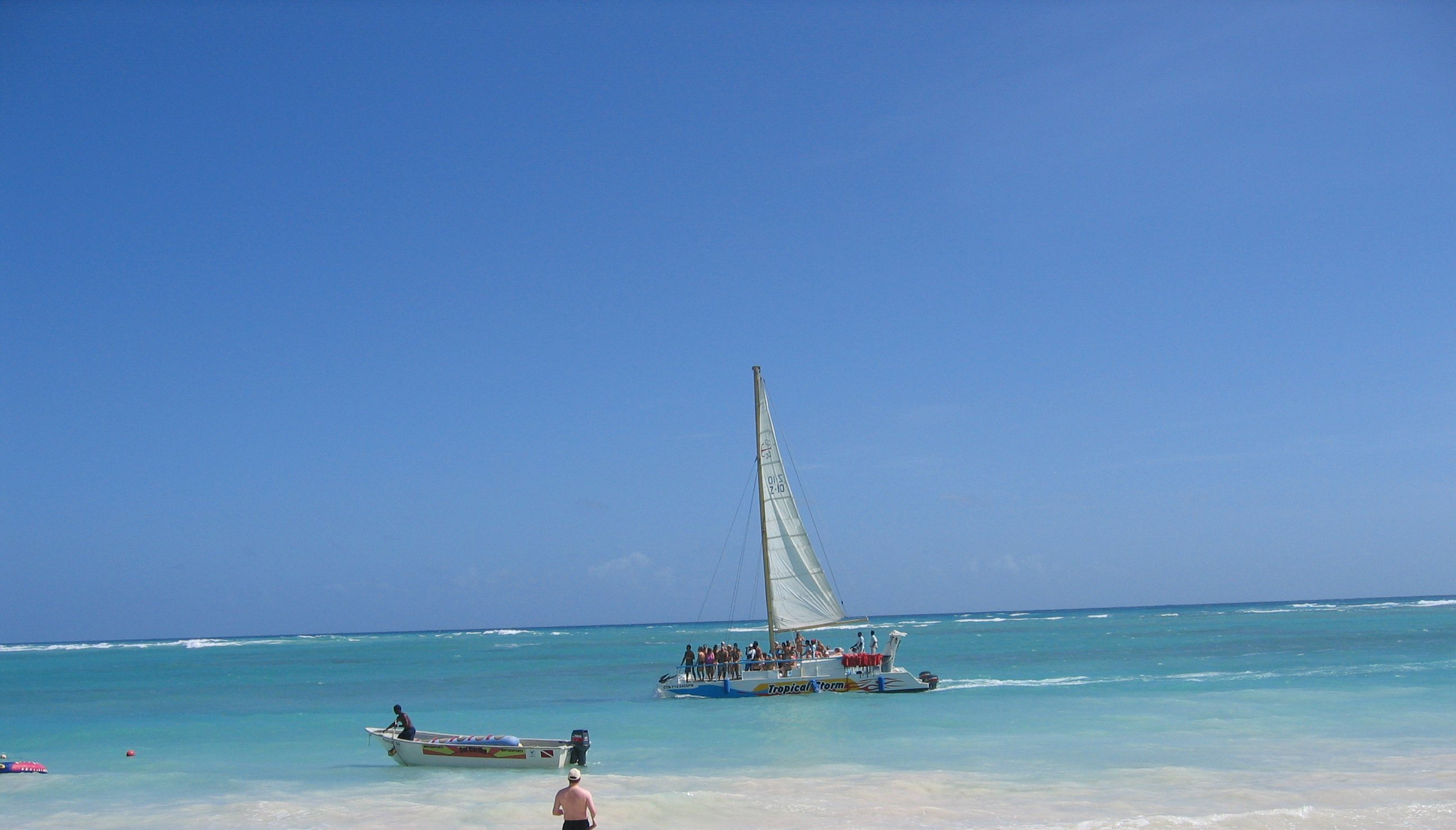 Playa Bávaro desde la orilla: agua turquesa de tres tonos, un catamarán de excursión saliendo y una lancha fondeada. Al fondo se ve la línea de rompiente del arrecife.